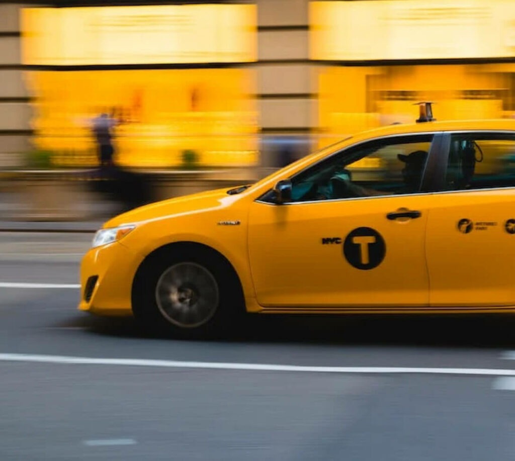 Een gele taxi rijdt door een straat, met gebouwen en voetgangers op de achtergrond.